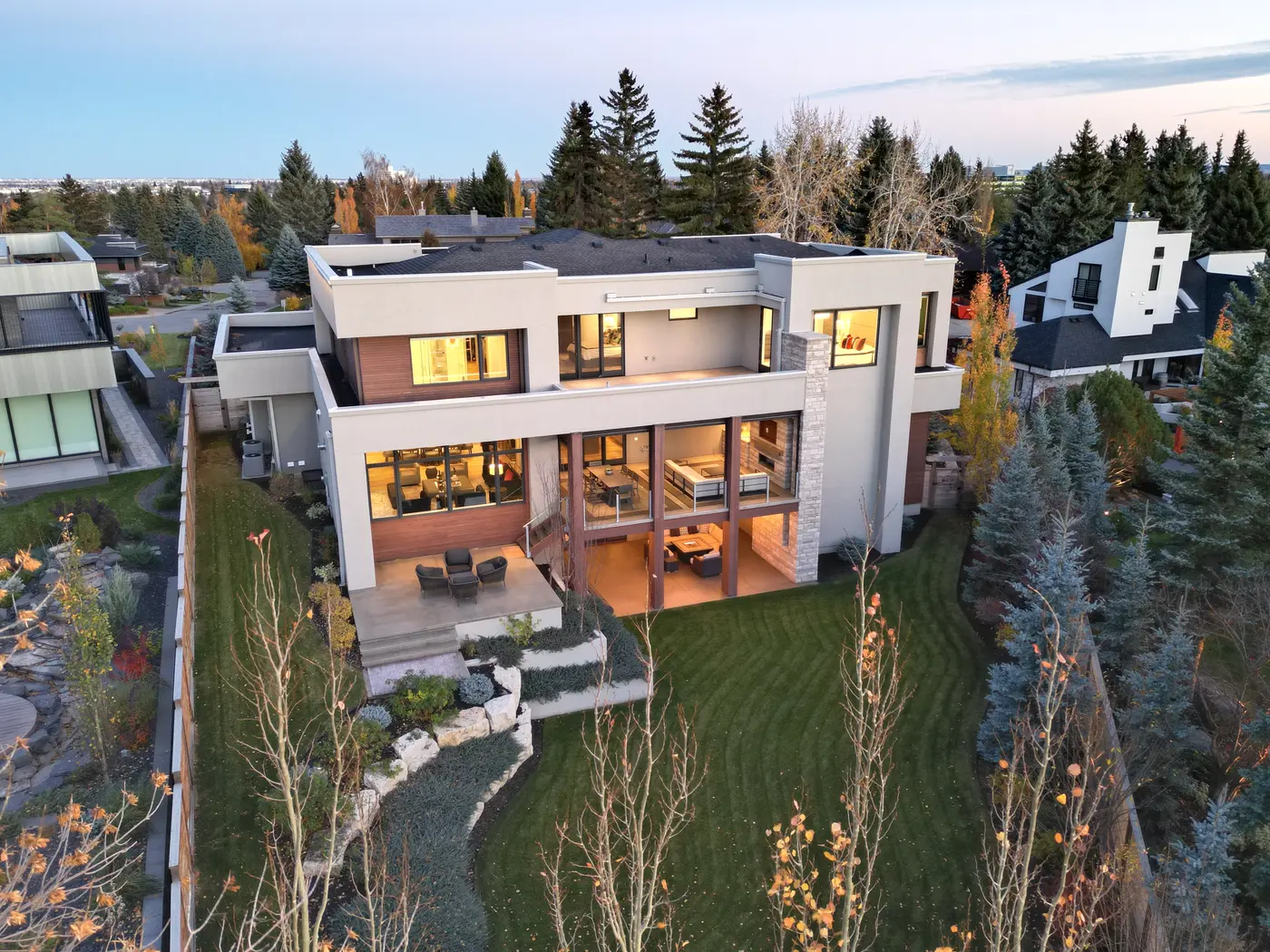 A twilight aerial of the rear facade captures the indoor-outdoor flow, from the glowing great room and covered fireplace lounge to the balcony off the primary suite.